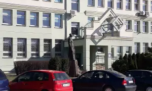 Pomnik przed budynkiem z oknami i kratami. Obok czerwony samochód, a dalej ciemny. KN rejestracja na tablicy.