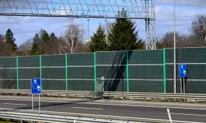 Znak informuje o odcinkowym pomiarze prędkości na A1. Za zieloną barierą dźwiękochłonną widać drzewa i niebo.