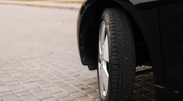Błyszczące, czarne auto na kostce brukowej. Widoczna opona zimowa, której wiek można ocenić po bieżniku.