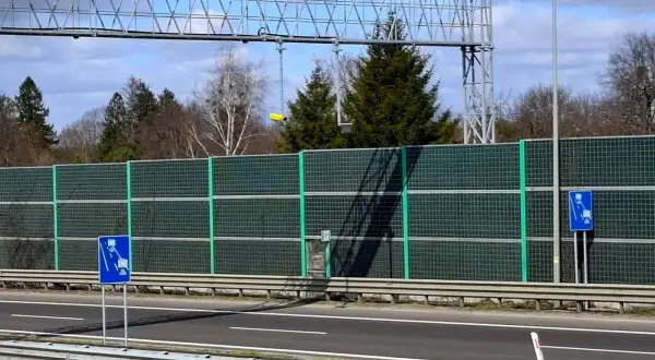 Znak informuje o odcinkowym pomiarze prędkości na A1. Za zieloną barierą dźwiękochłonną widać drzewa i niebo.
