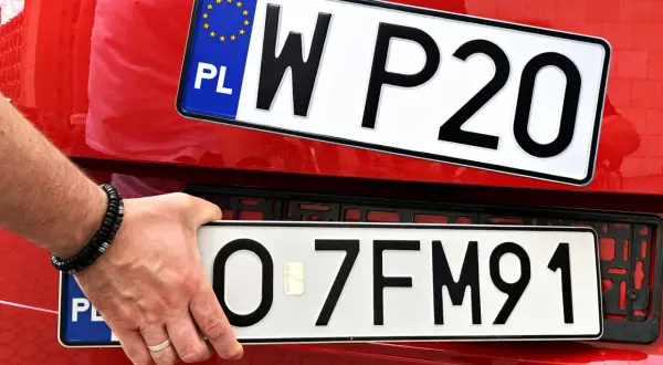 Ręka trzyma tablicę rejestracyjną z napisem "O 7FM91". Obok widać inną tablicę "W P20". To początek ep rejestracja.