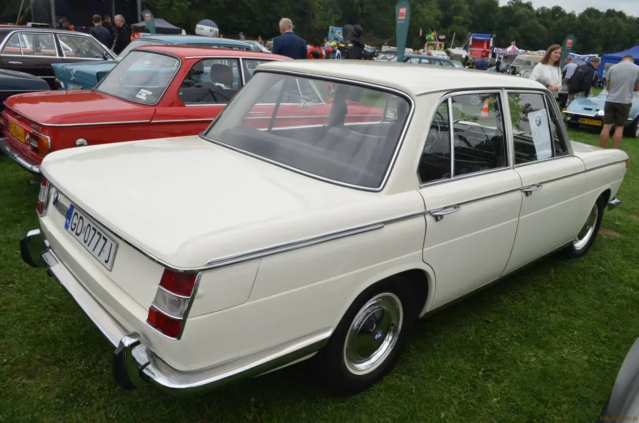 Białe BMW E10 z lat 70. prezentuje się elegancko na zielonej trawie, obok czerwonego klasyka.