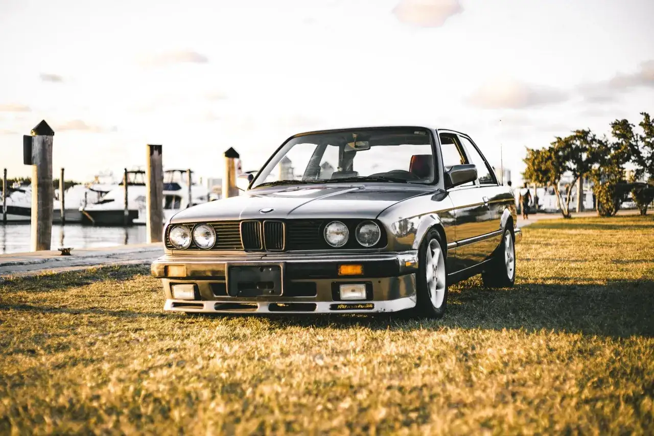 Klasyczne bmw e10 na trawie przy nabrzeżu, z łodziami w tle.