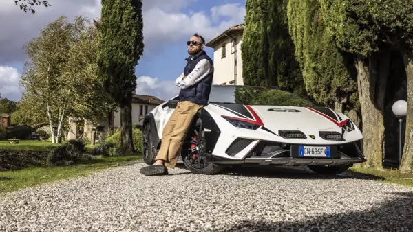 Mężczyzna opiera się o białe Lamborghini Sterrato z czerwono-czarnymi akcentami, zaparkowane na żwirowej drodze wśród drzew.