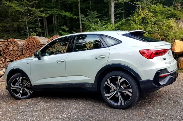 Nowy Audi Q3 2025 w kolorze białym, zaparkowany na leśnej drodze, z widocznymi stosami drewna w tle.
