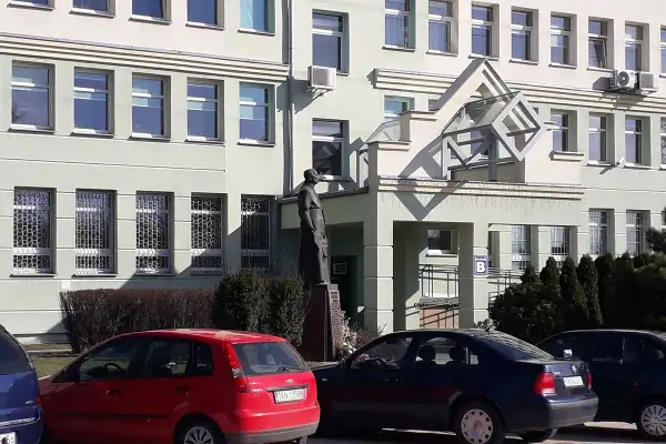 Pomnik przed budynkiem z oknami i kratami. Obok czerwony samochód, a dalej ciemny. KN rejestracja na tablicy.