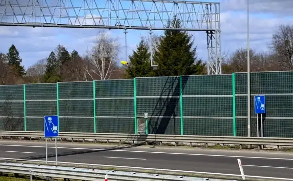 Znak informuje o odcinkowym pomiarze prędkości na A1. Za zieloną barierą dźwiękochłonną widać drzewa i niebo.