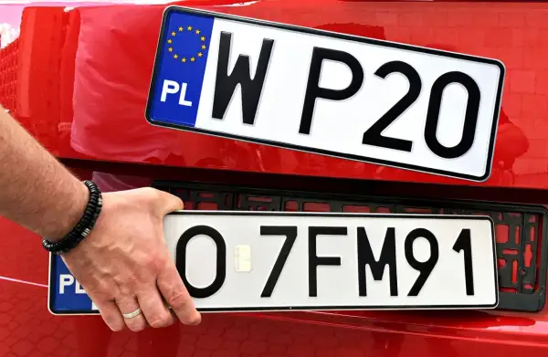 Ręka trzyma tablicę rejestracyjną z napisem "O 7FM91". Obok widać inną tablicę "W P20". To początek ep rejestracja.